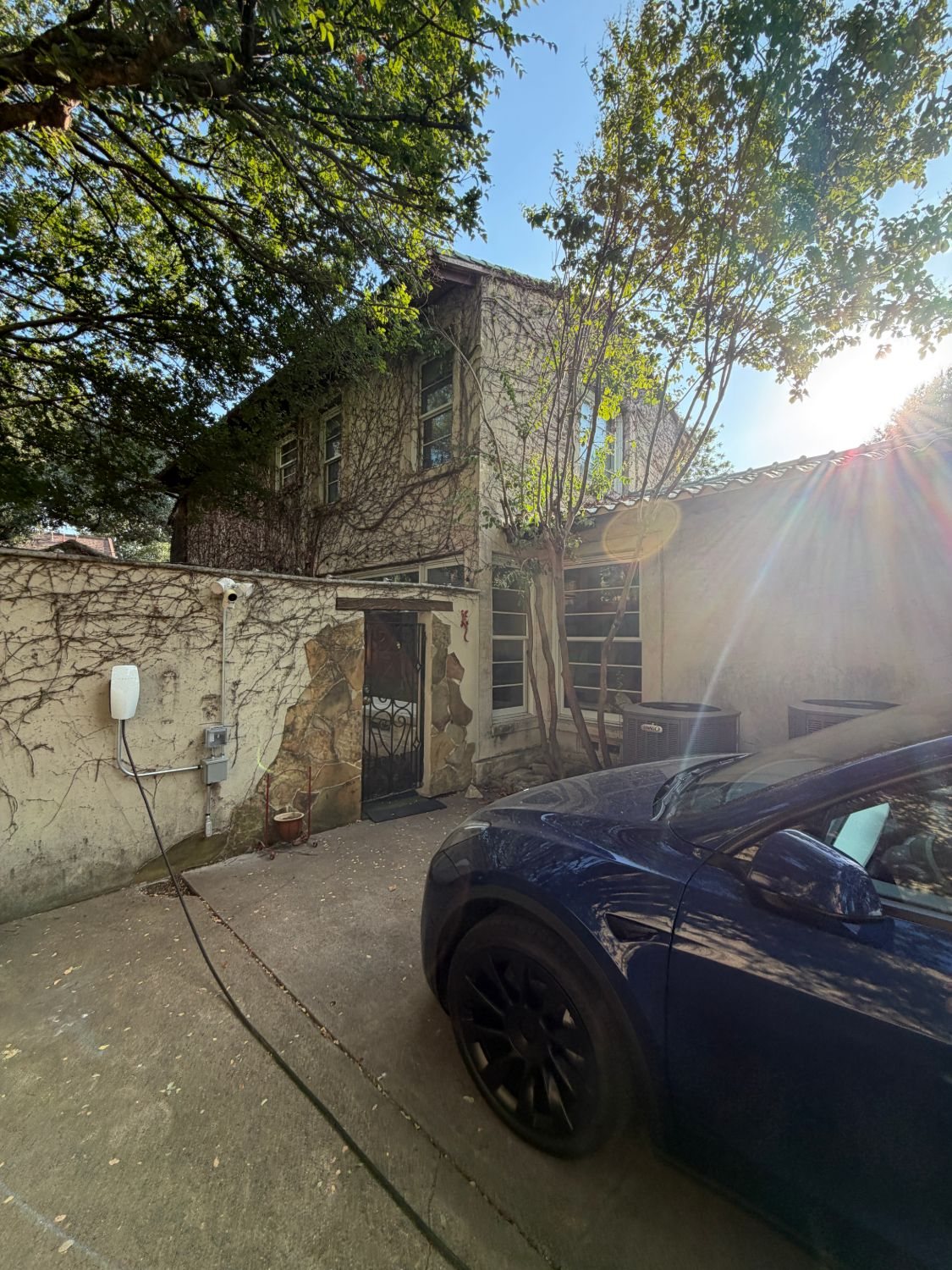 A Tesla Wall Connector on a stucco wall, with its cable running to a car in the driveway